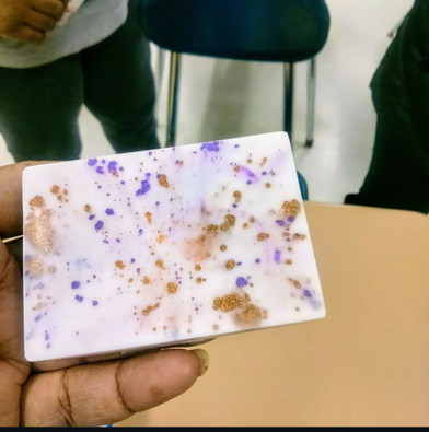 Hand holding a decorative bar of soap with purple and gold speckles on a blurred background