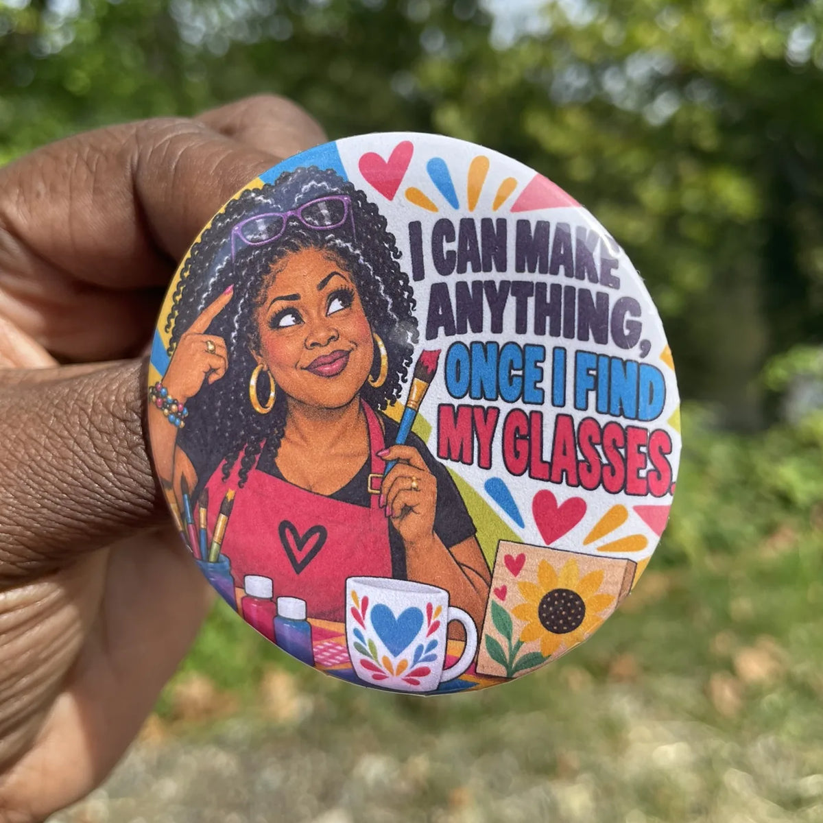 A hand holds a round pin-back button badge featuring a colorful illustration of a woman painting with the text 'I CAN MAKE ANYTHING, ONCE I FIND MY GLASSES.' The background is a blurred outdoor green setting.