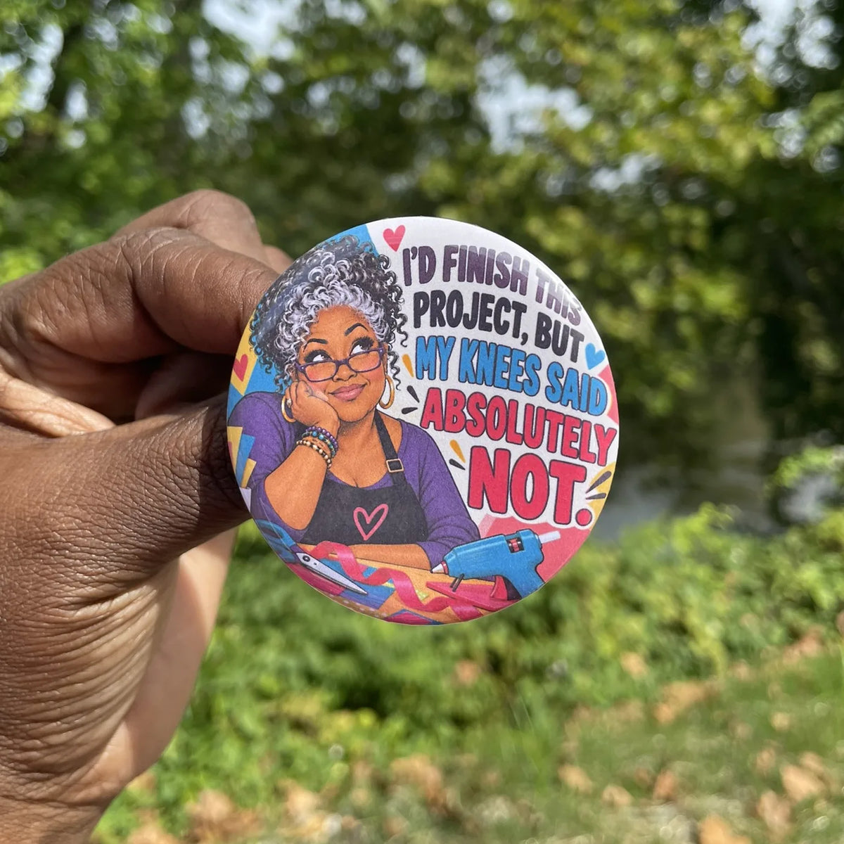 Hand holding a round button with an illustrated older woman with silver hair and crafting supplies and the words I'd Finish This Project, But My Knees Said Absolutely Not.