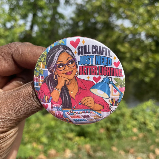 Hand holding a round button with an illustrated crafter in purple glasses stringing beads under a blue lamp and the words Still Crafty. Just Need Better Lighting.